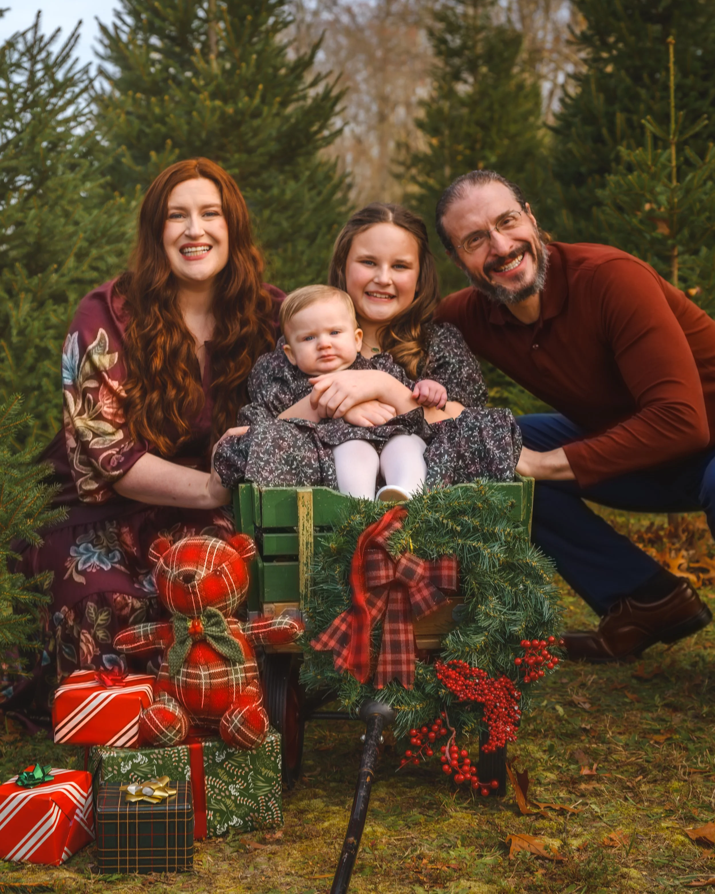A family of four, including a man, woman, teenage girl, and baby, smiling outdoors among Christmas trees, surrounded by Christmas decorations and wrapped gifts.