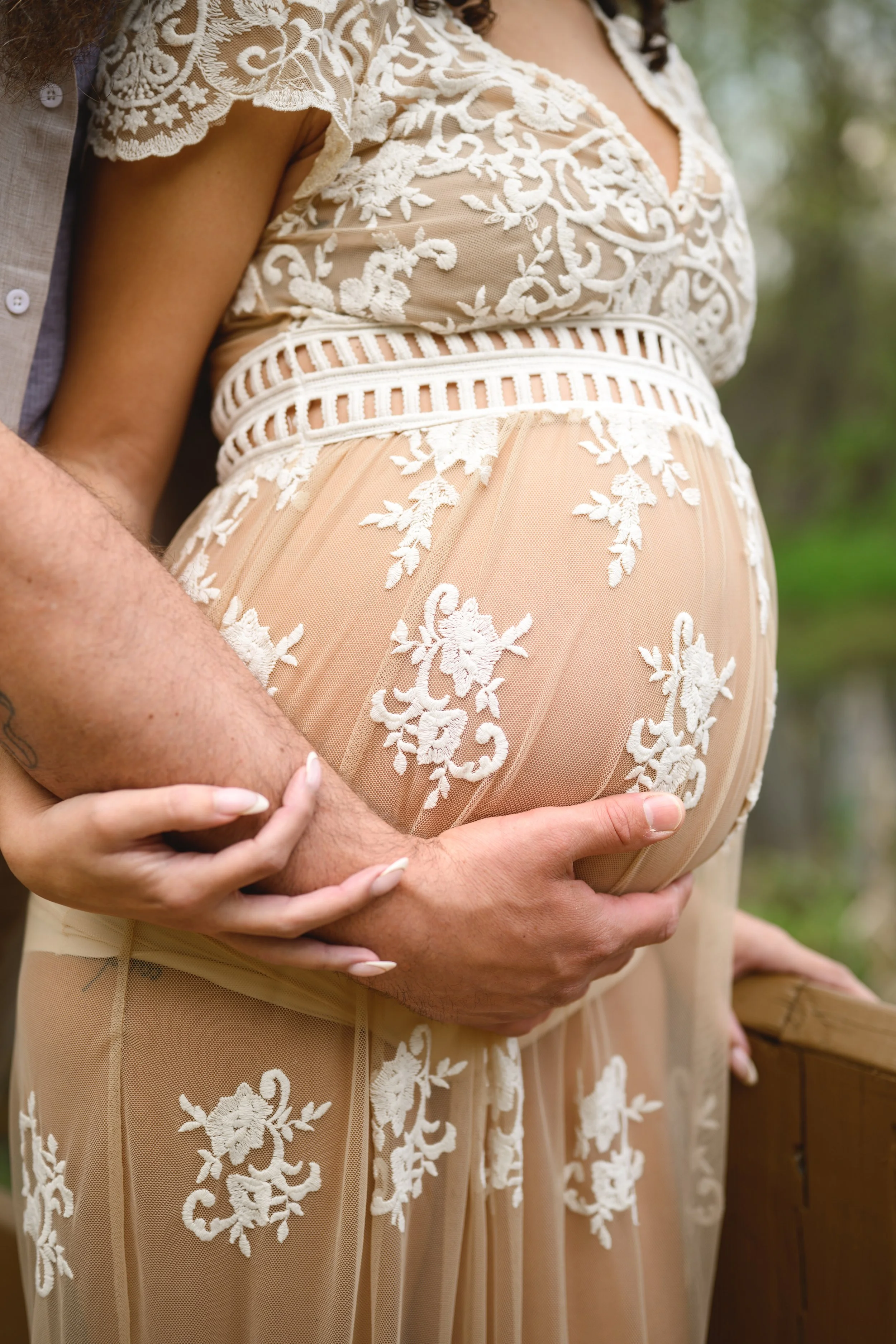 Close-up of a pregnant woman in a beige lace dress with floral embroidery, being gently held from behind by a person with an arm visible.