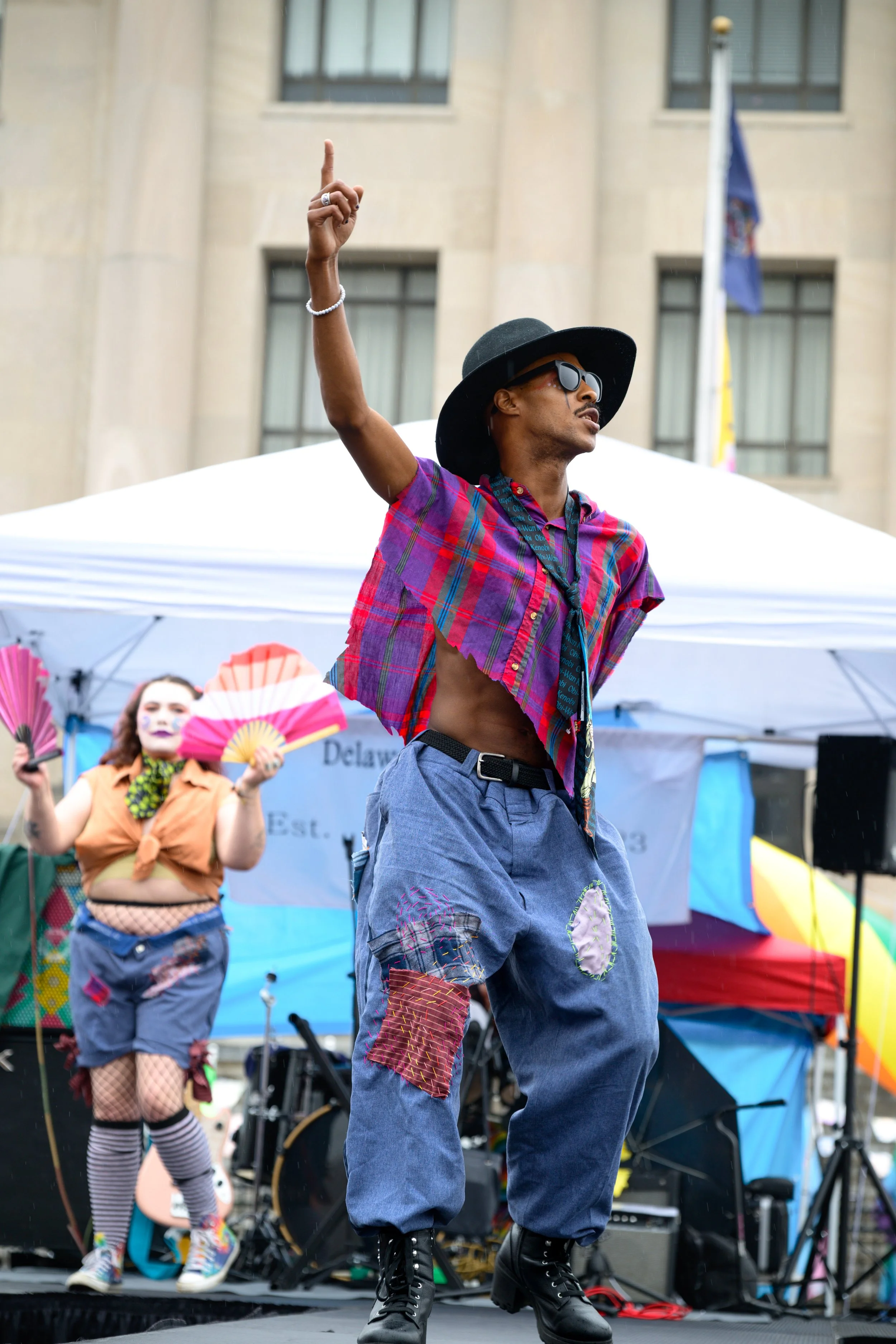 A person dancing on stage at a pride event, wearing a black wide-brimmed hat, sunglasses, a colorful cropped shirt, baggy jeans with colorful patches, and black combat boots, with a person in drag in the background holding a pink fan.
