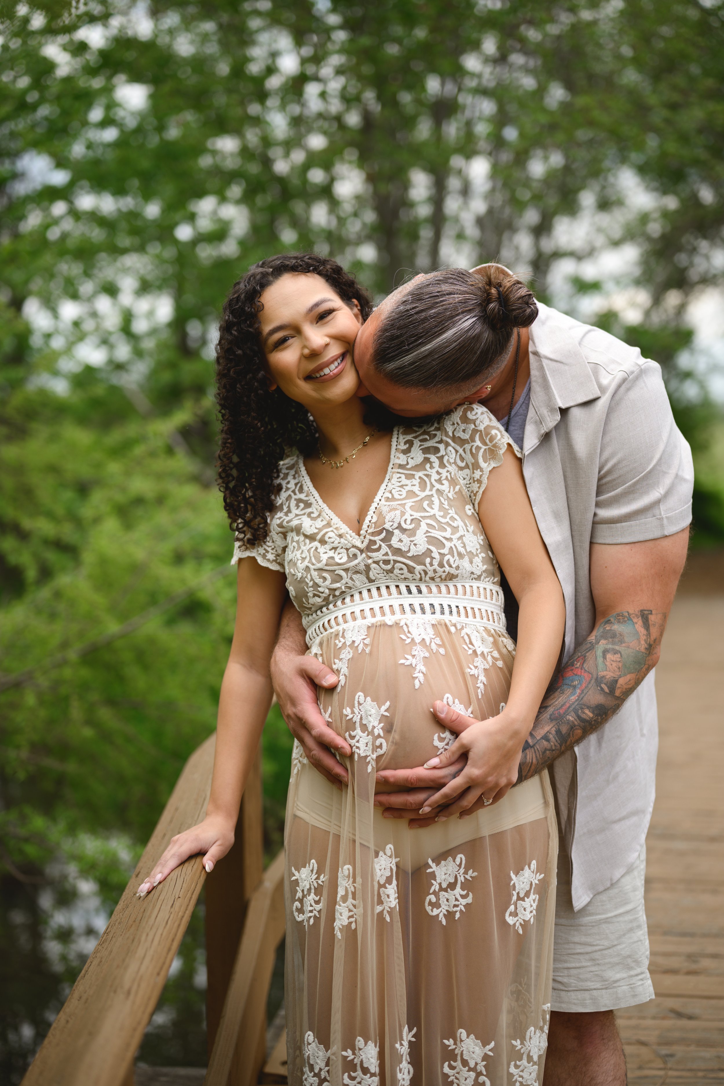 A smiling woman with curly hair in a lace dress stands on a wooden bridge, while a man with tattoos and a bun kisses her cheek, both holding her pregnant belly. The scene is outdoors with green trees in the background.