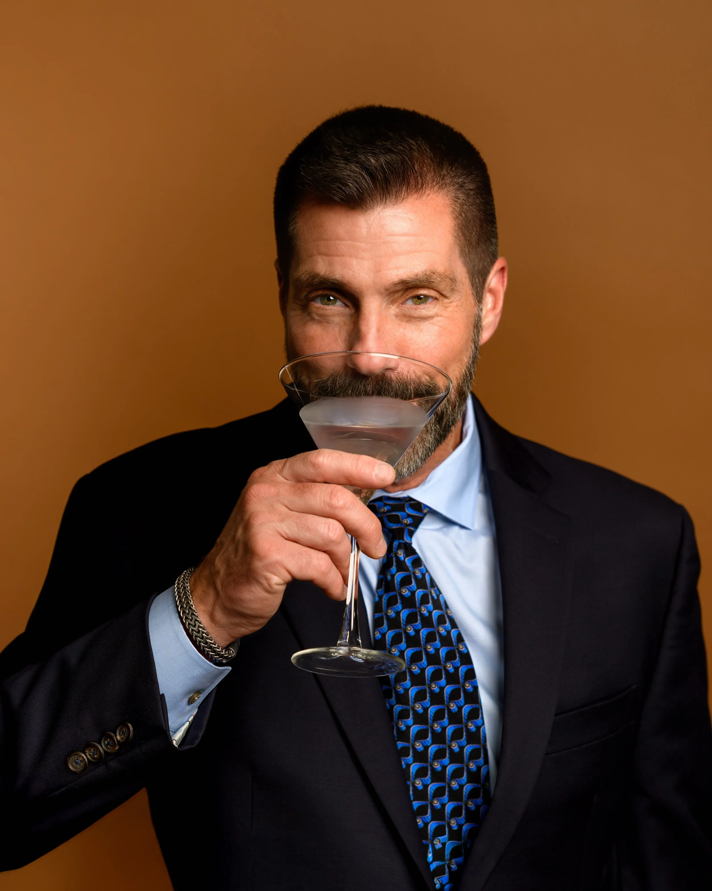A headshot of a man with dark hair and a beard, dressed in a suit and tie, holding a martini glass up to his lips.