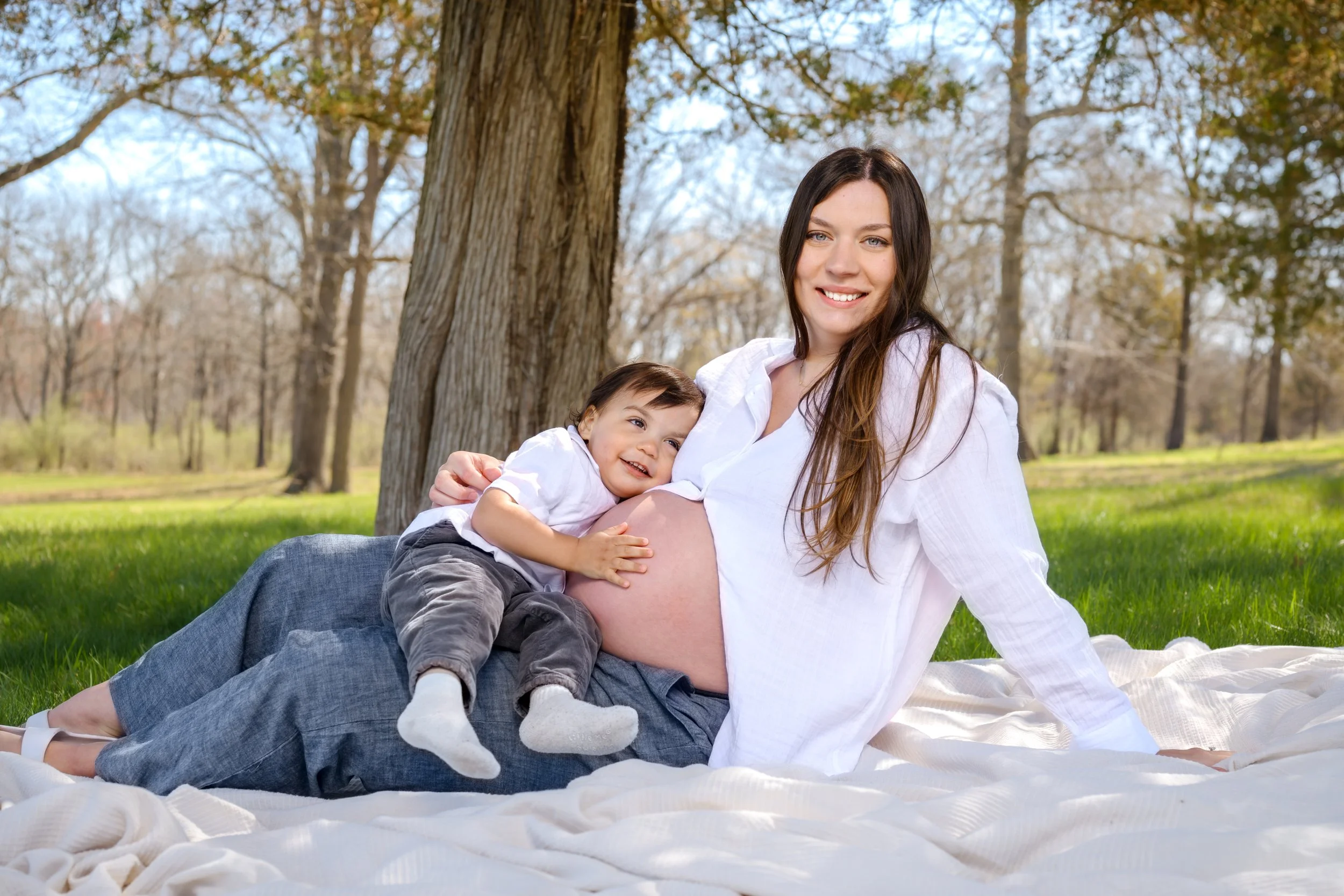 A pregnant woman with long brown hair, wearing a white shirt, sitting on a blanket in a park with trees in the background, smiling as she hugs a young boy.