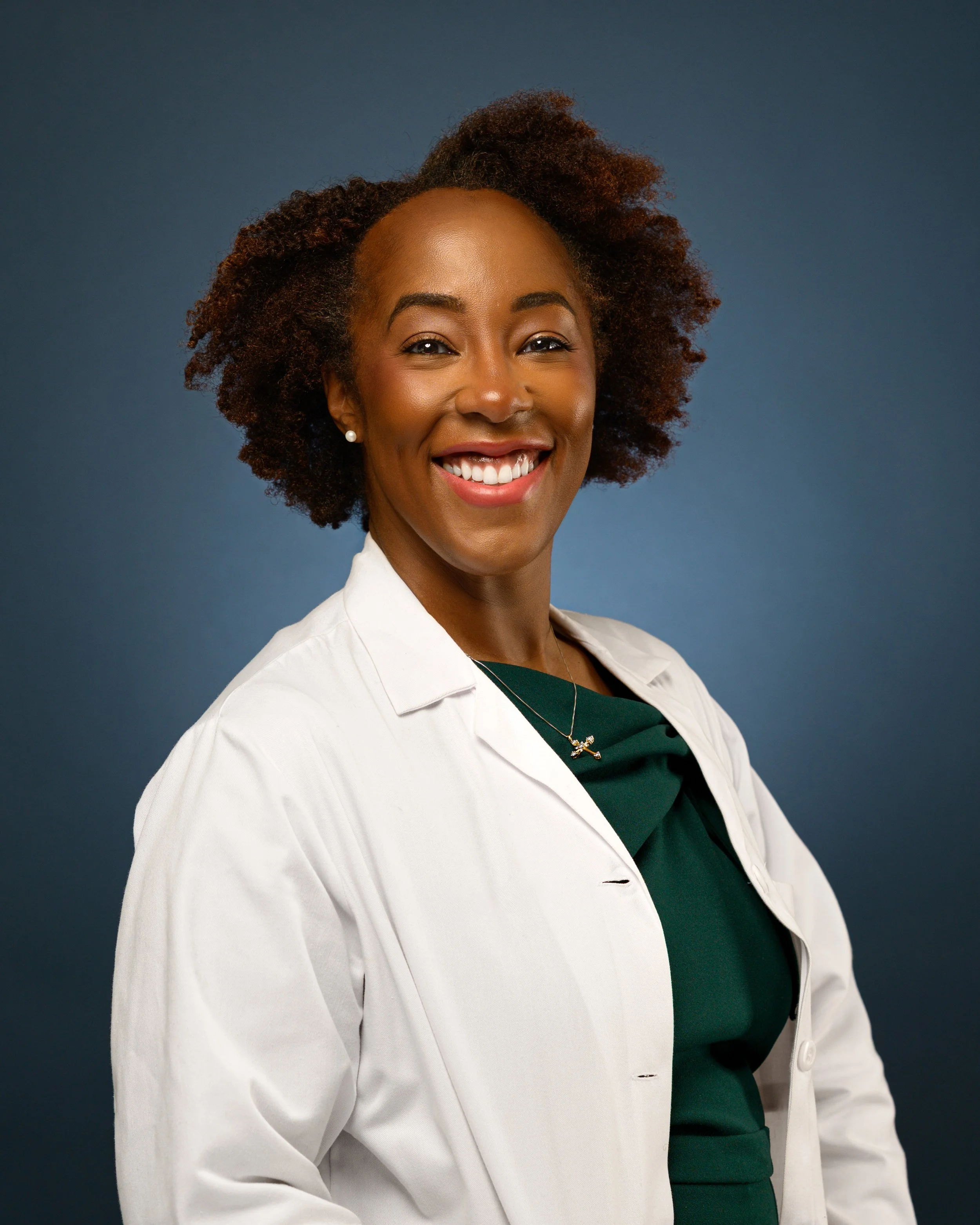 A headshot of smiling woman with natural curly hair wearing a white doctor's coat over a dark green top, standing against a blue background.