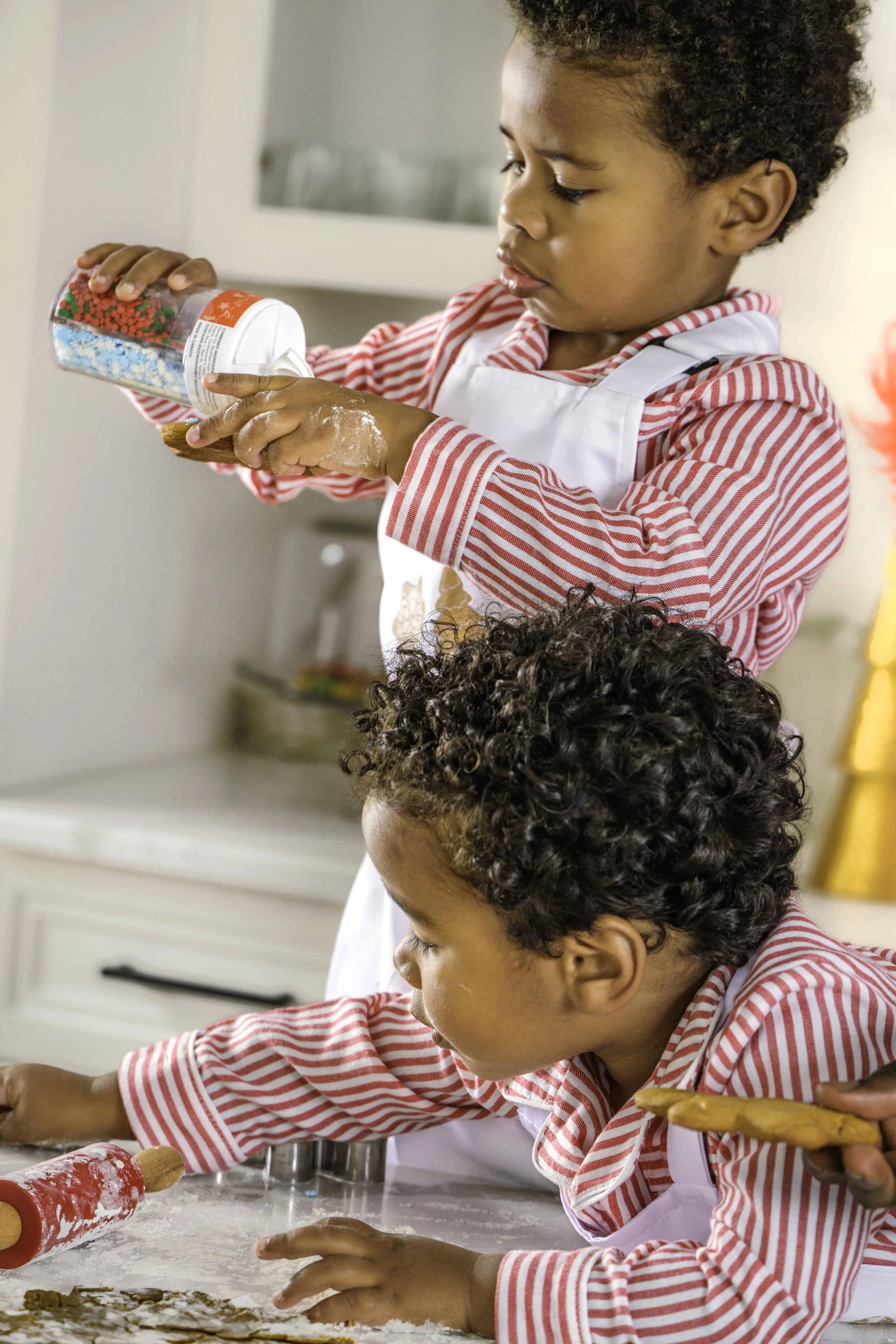 Two children baking cookies in a kitchen. One child is pouring colorful baking sprinkles onto cookie dough while the other is reaching for a cookie cutter. Both are wearing red and white striped shirts and aprons.