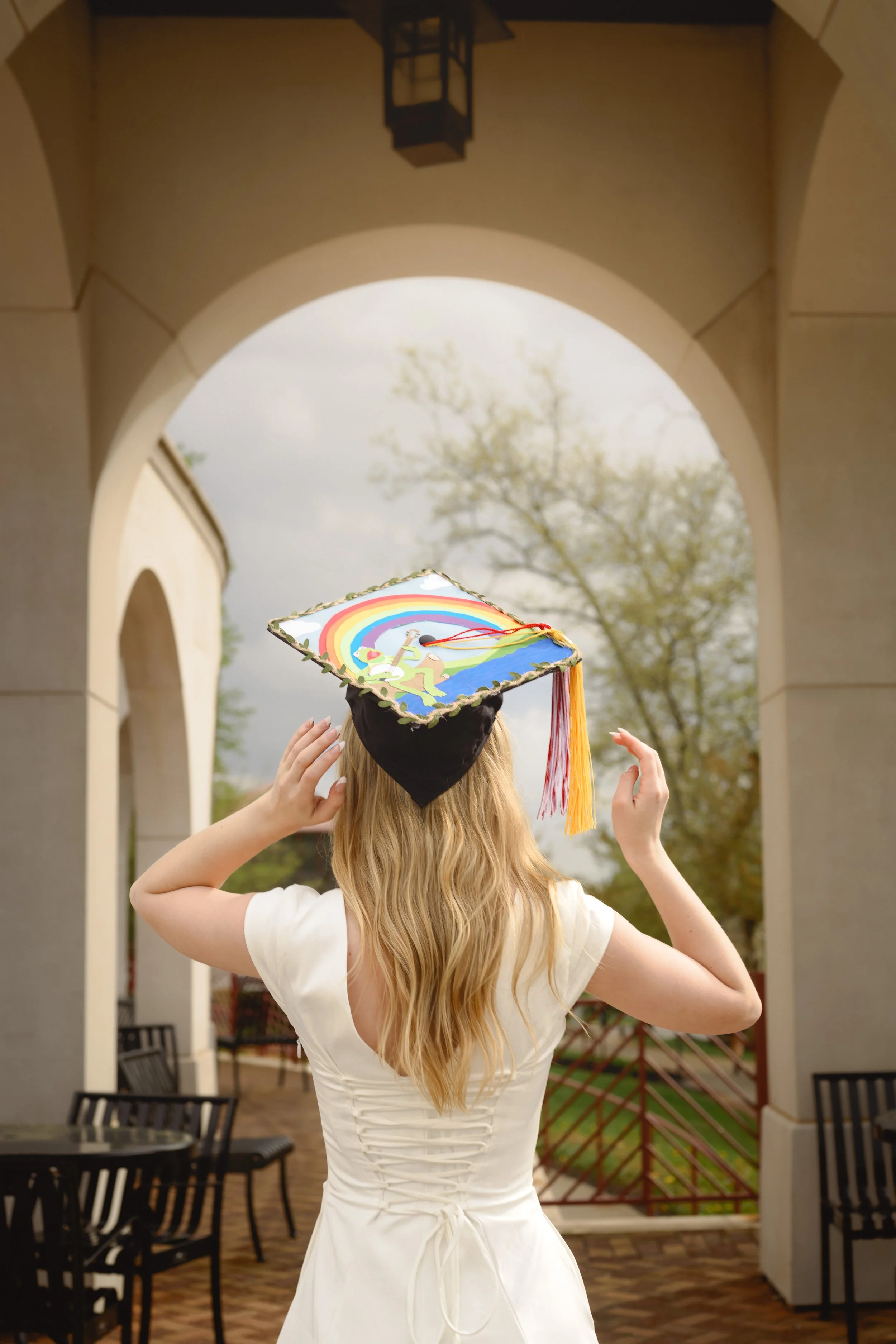 A woman with long blonde hair is seen from behind, wearing a white dress and a black face mask, adjusting her aquamarine graduation cap with rainbow graphics as she stands in an outdoor corridor with beige walls and a large archway. Trees are visible in the background.