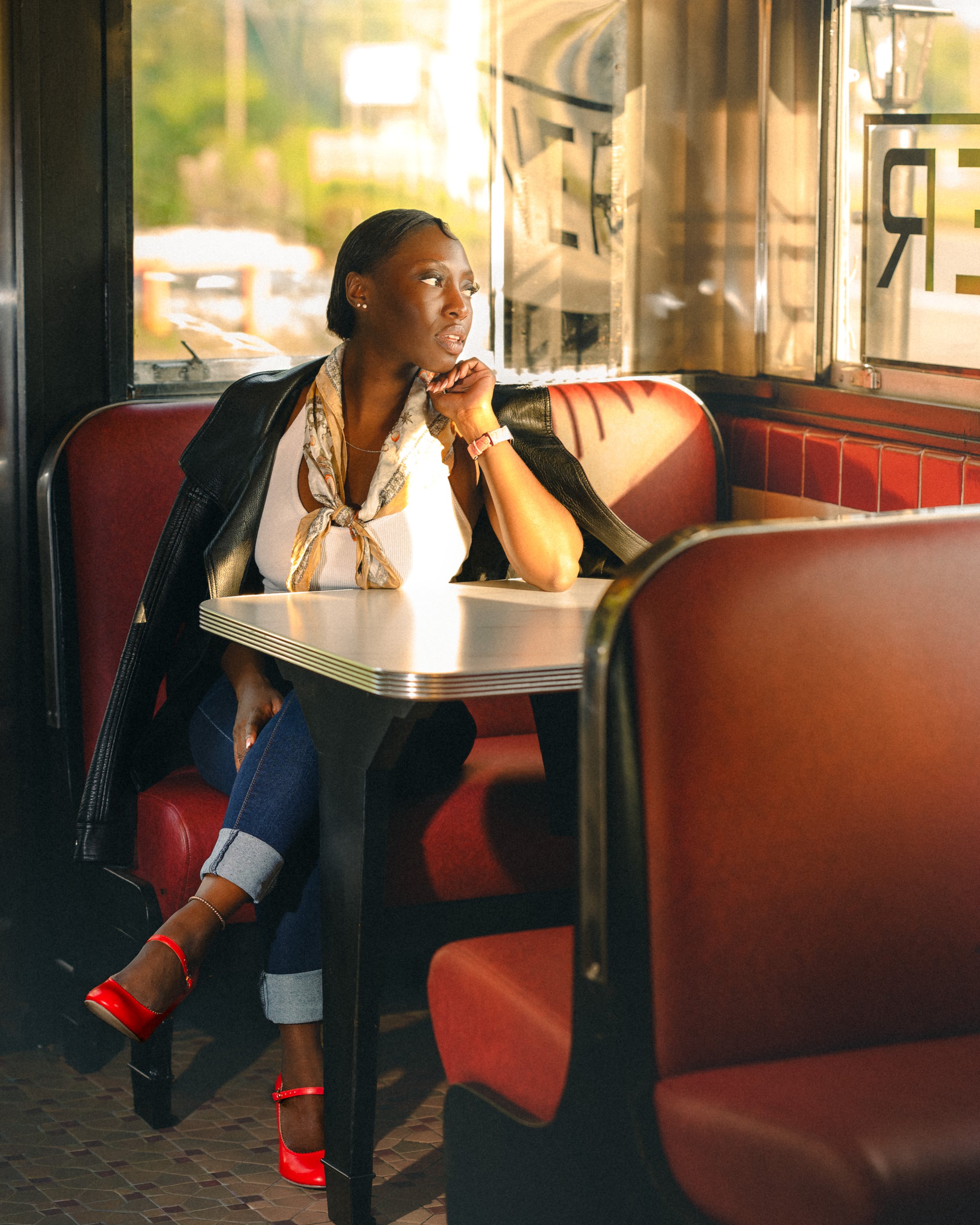 A woman in a leather jacket sitting alone at a booth in a diner, looking out the window as the setting sunlight streams in.