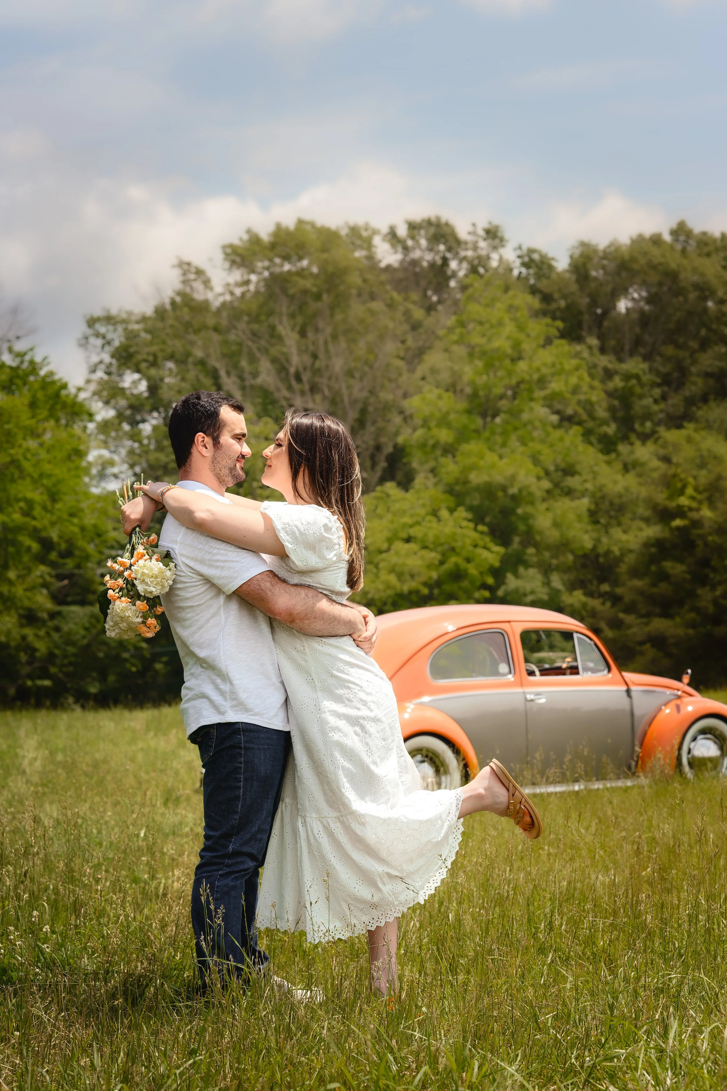 A couple embracing in a field with a vintage orange and gray car in the background.