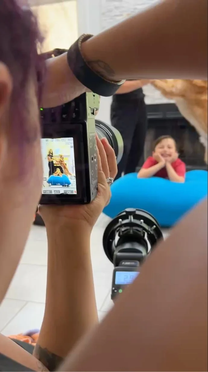 Photographer taking a picture of a young girl lying on the floor in a living room, with a cheerful and relaxed expression, wearing a red shirt, seen through the viewfinder of the camera.