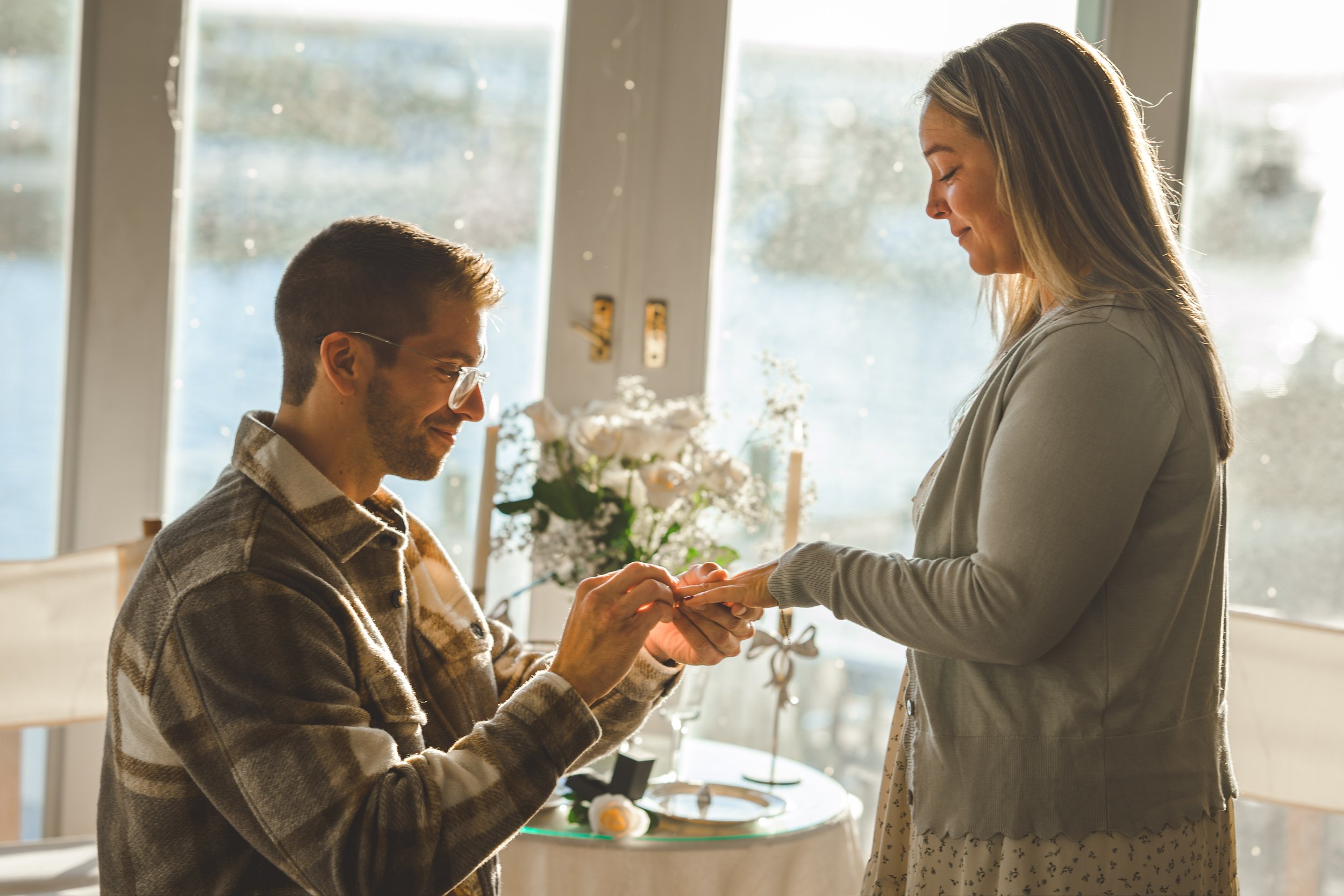 A man is proposing marriage to a woman in a bright room with large windows, with sunlight streaming in. The man is kneeling and placing a ring on the woman's finger. The woman looks at the man with a smile. There are flowers and a small table in the background.