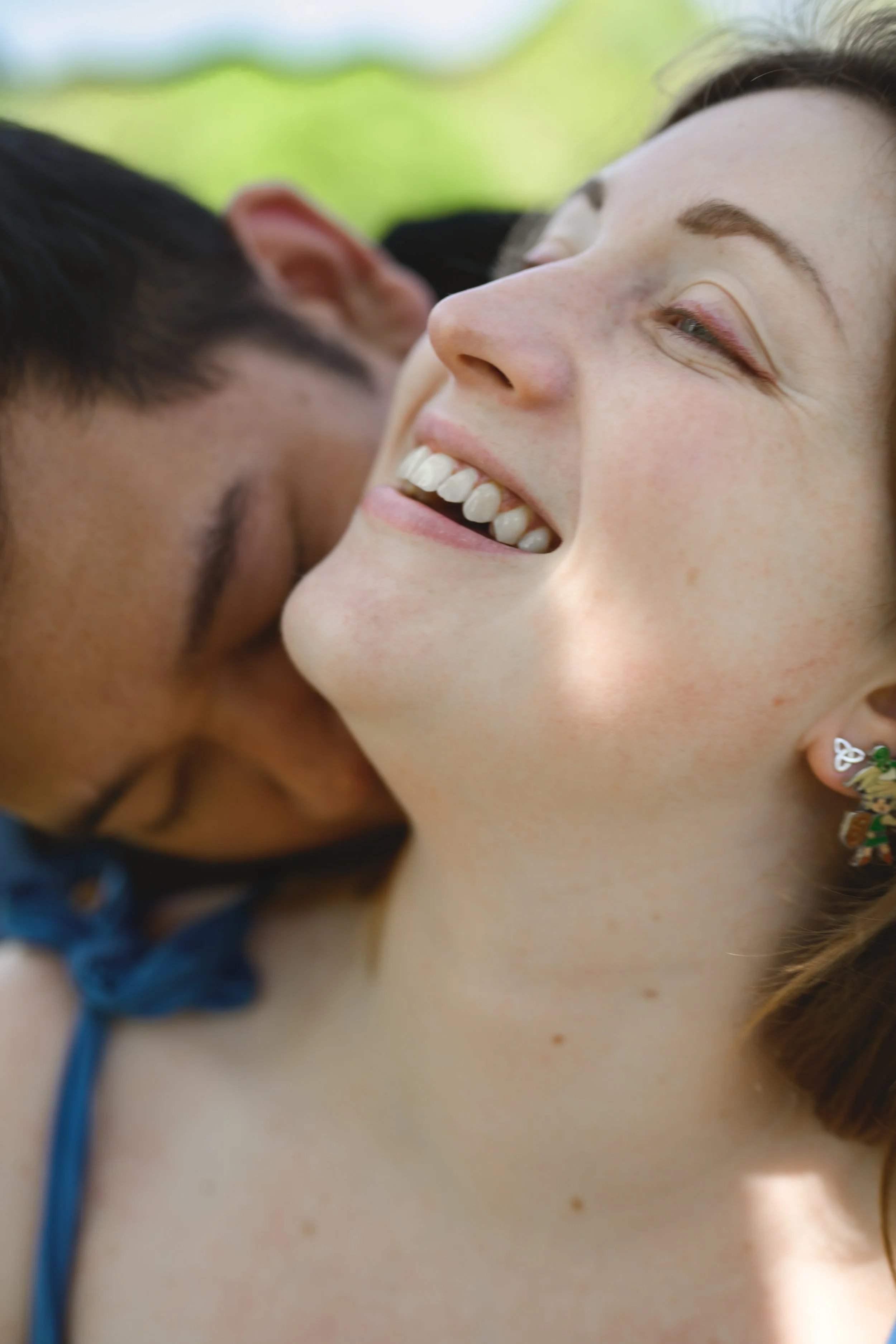 Close-up of a happy young woman with brown hair, smiling, with her eyes closed, outdoors with a blurred green background.