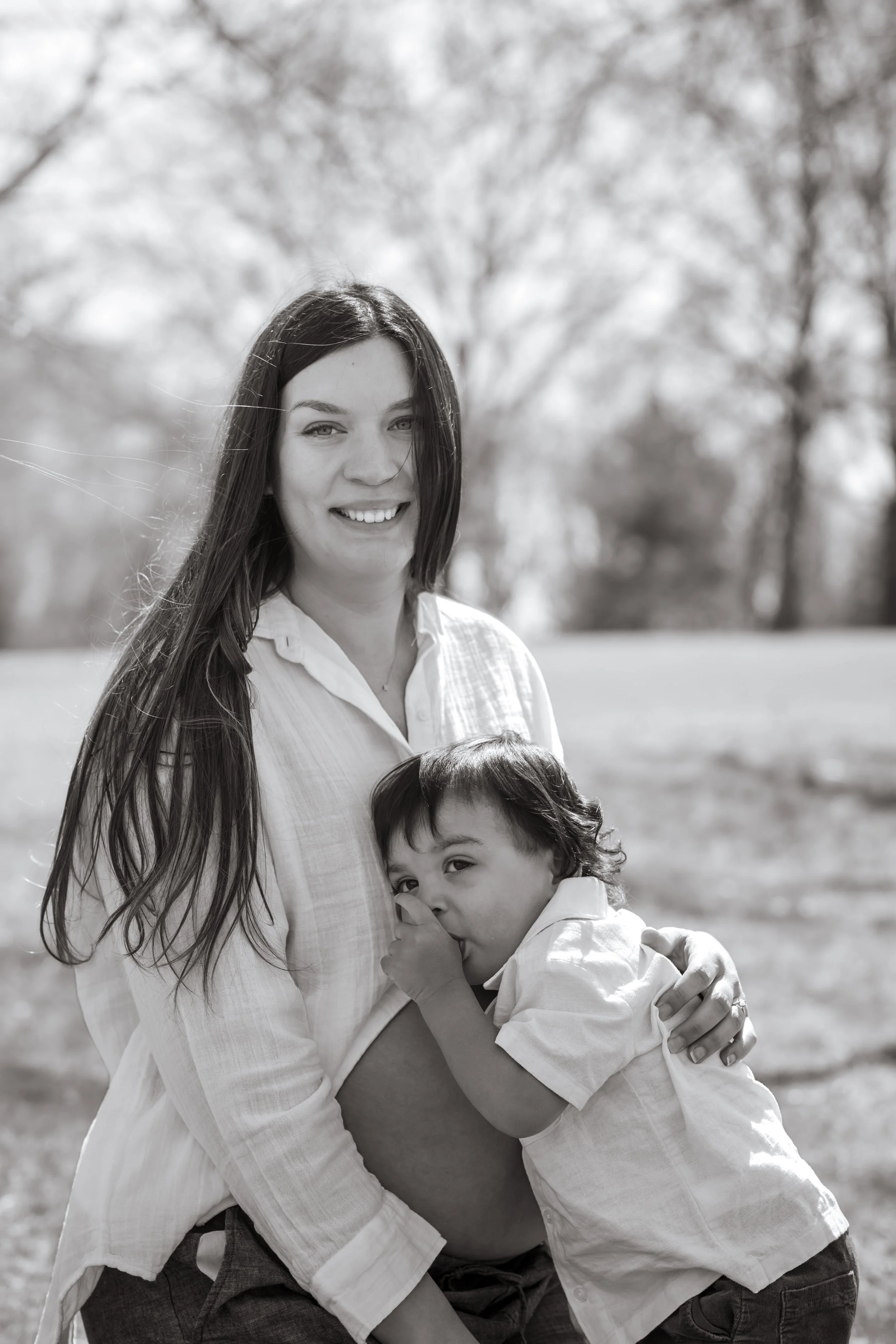 A pregnant woman with long dark hair smiling, holding a young child in her arms outdoors. The child has dark, curly hair and is biting his finger.