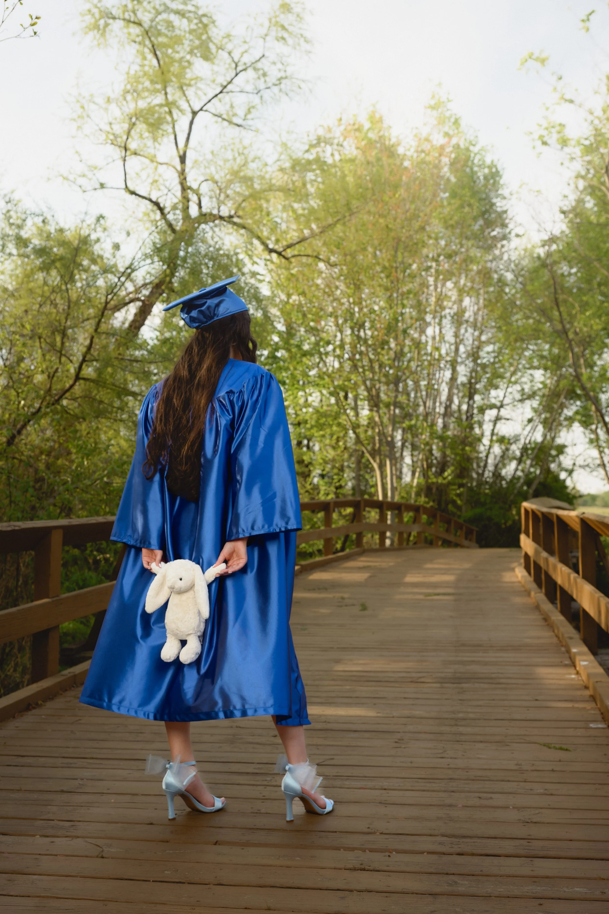 A woman in a blue graduation gown and cap holding a stuffed bunny toy on a wooden bridge surrounded by trees.