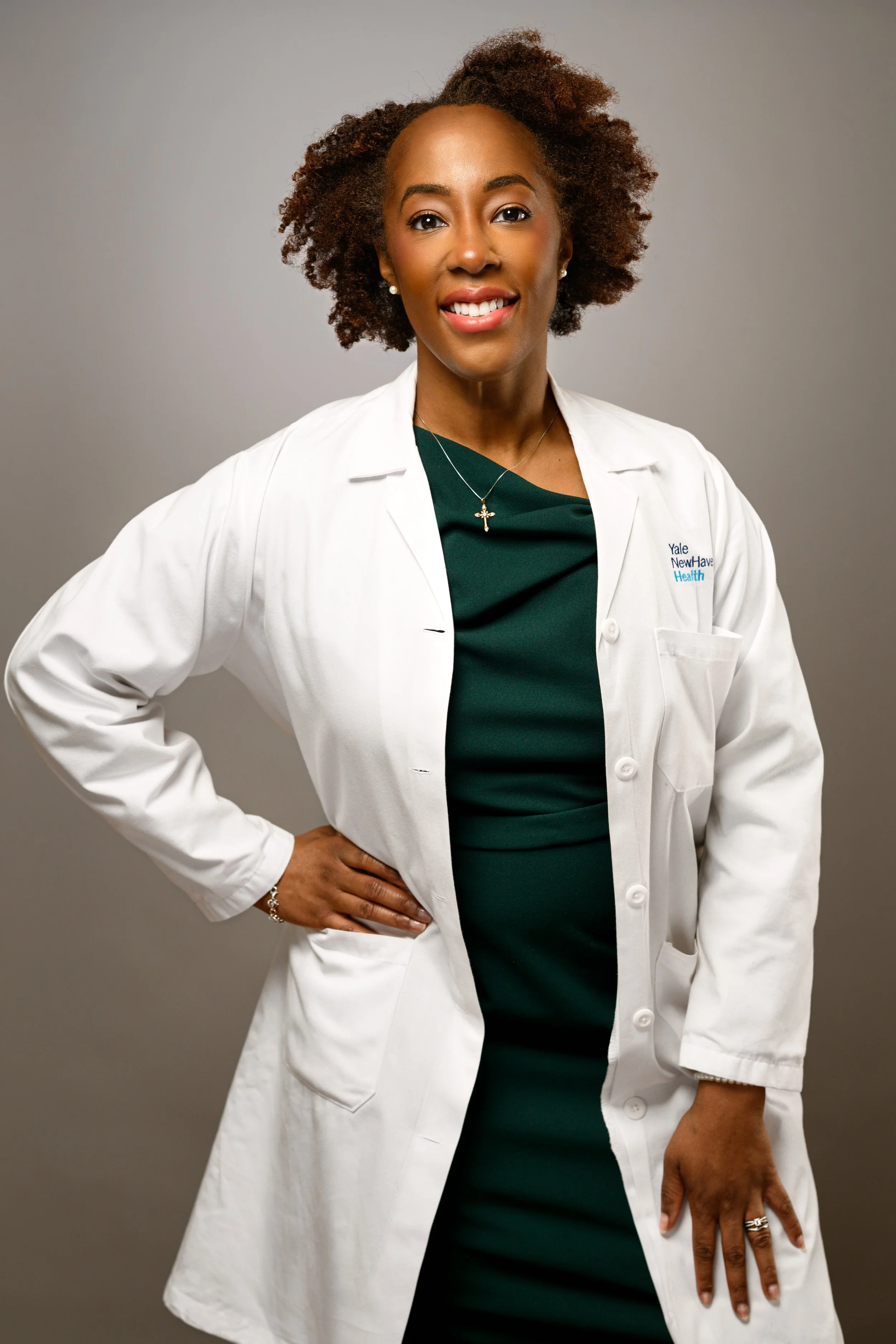A headshot of a woman with natural curly hair wearing a green dress and a white lab coat, standing confidently with one hand on her hip, smiling at the camera against a neutral background.