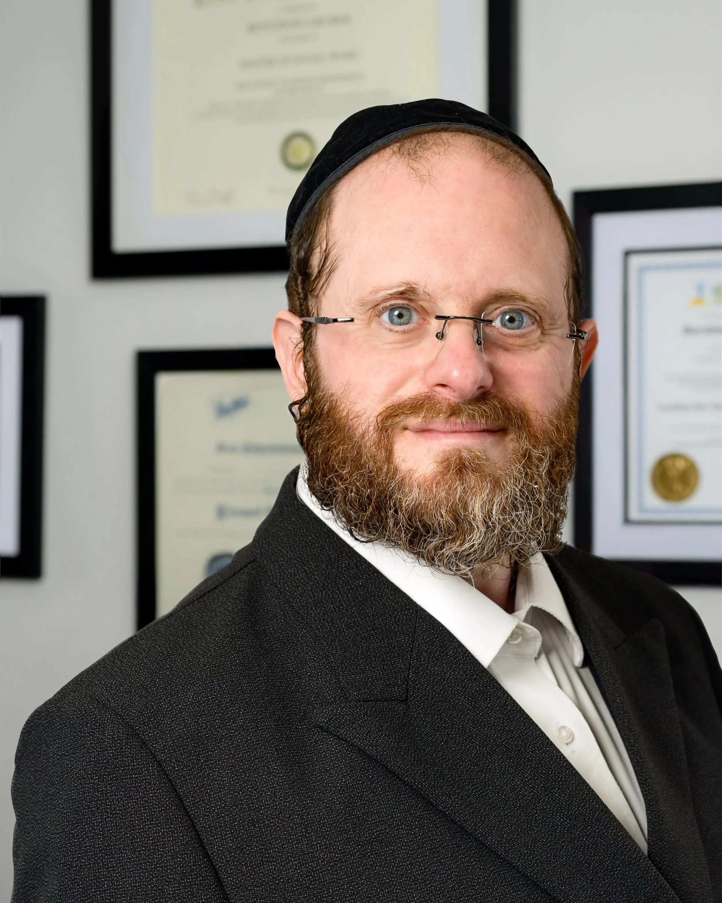 Headshot of a Jewish man with a beard and glasses, wearing a black suit and a kippah, standing in front of framed certificates or diplomas.