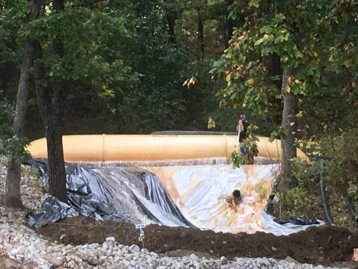 Person standing near the top of a half-buried pool of water, which is covered with black and white plastic sheeting, surrounded by trees with fall foliage.