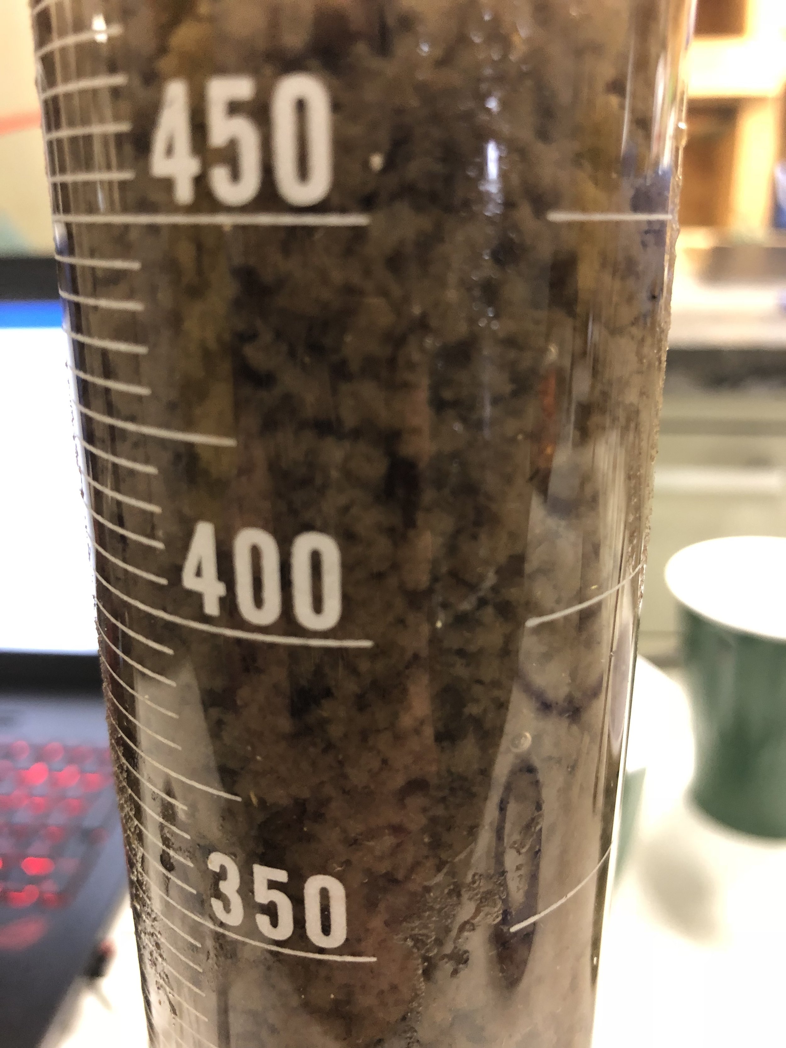 Close-up of a clear graduated measuring cup filled with a dark, muddy substance, with measurement markings from 350 to 450 milliliters visible.