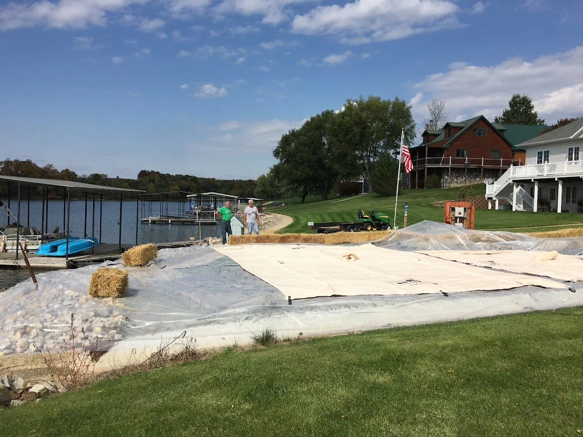 People working on a construction project near a lake, with houses, trees, and a cloudy sky in the background.