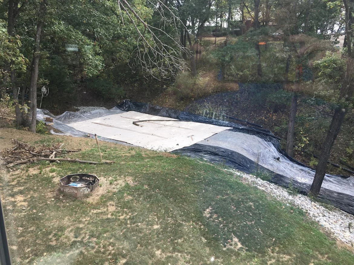 An outdoor scene with a partially covered muddy area, surrounded by trees and grass. There is a black plastic sheet and a white tarp covering part of the ground, with some rocks and debris nearby.
