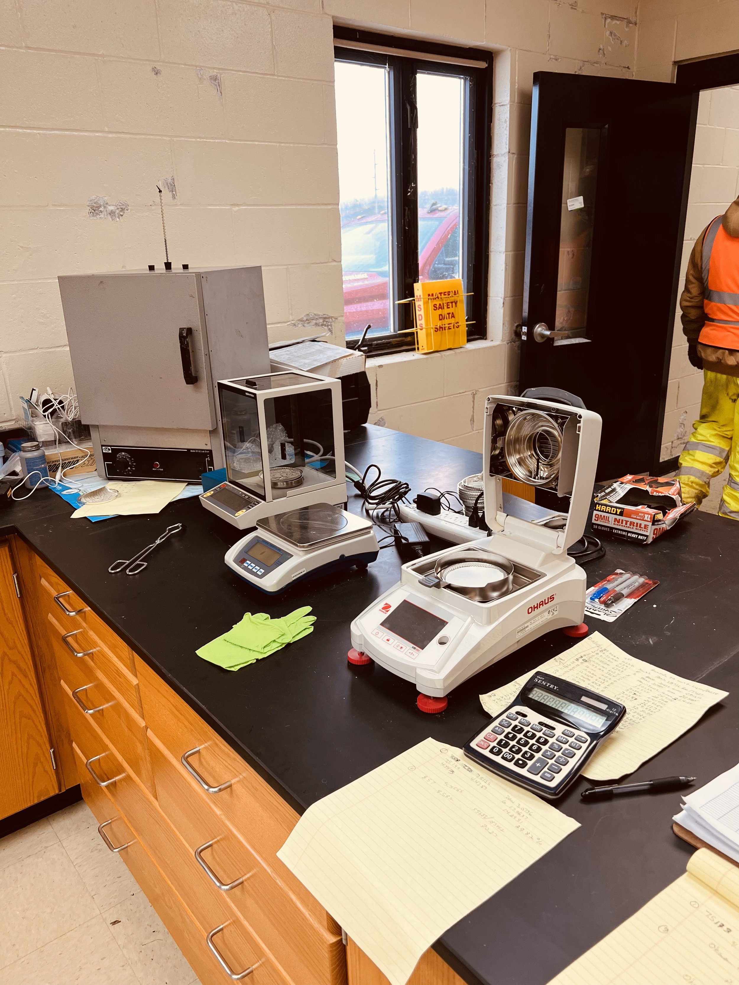 Laboratory workspace with scientific instruments, scales, a calculator, papers, gloves, and lab equipment on a black countertop, with a window and a partially visible person wearing a safety vest in the background.