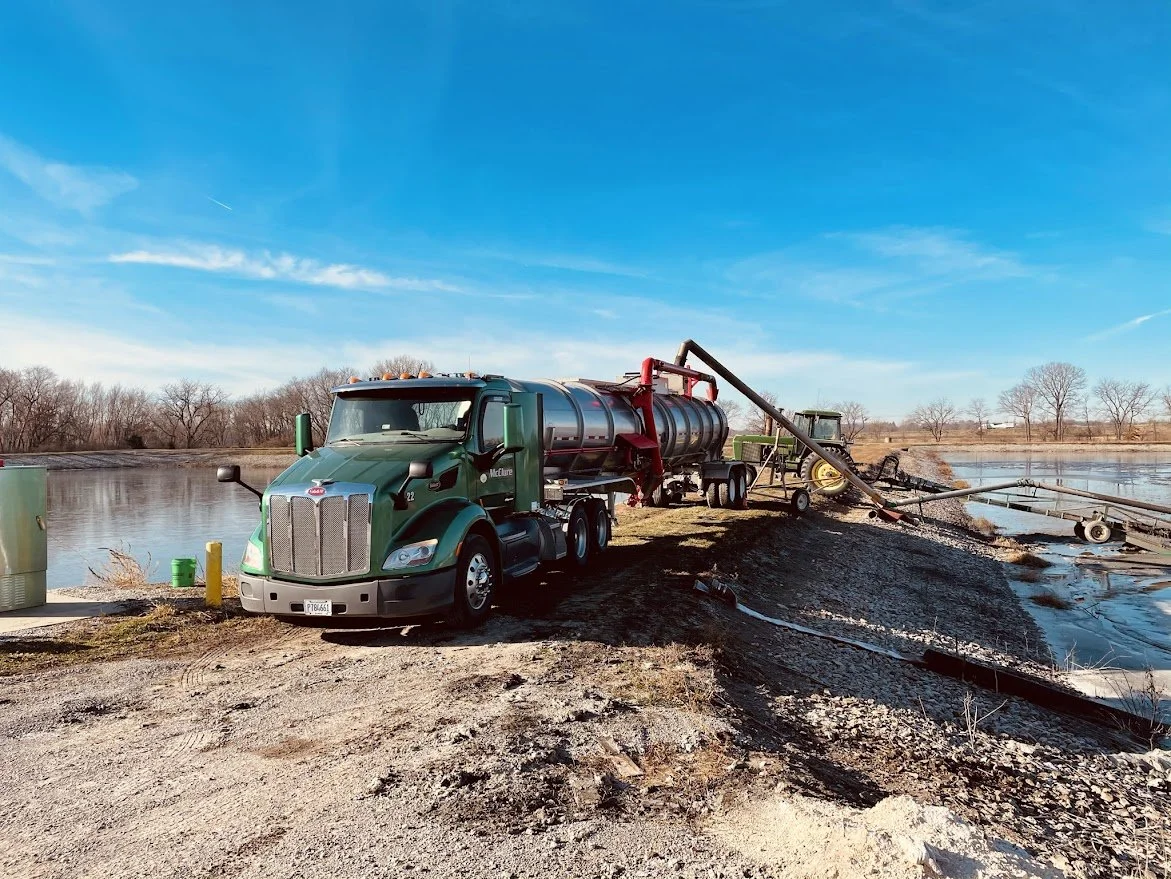 A large green tanker truck with a silver tank is parked near a body of water, connected to a pump system used for transferring liquids, with a tractor nearby and trees in the background under a clear blue sky.