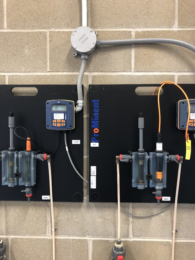 Industrial plumbing and filtration system with two black control panels labeled ProMinent, various tubes, filters, and electronic components mounted on a beige brick wall.