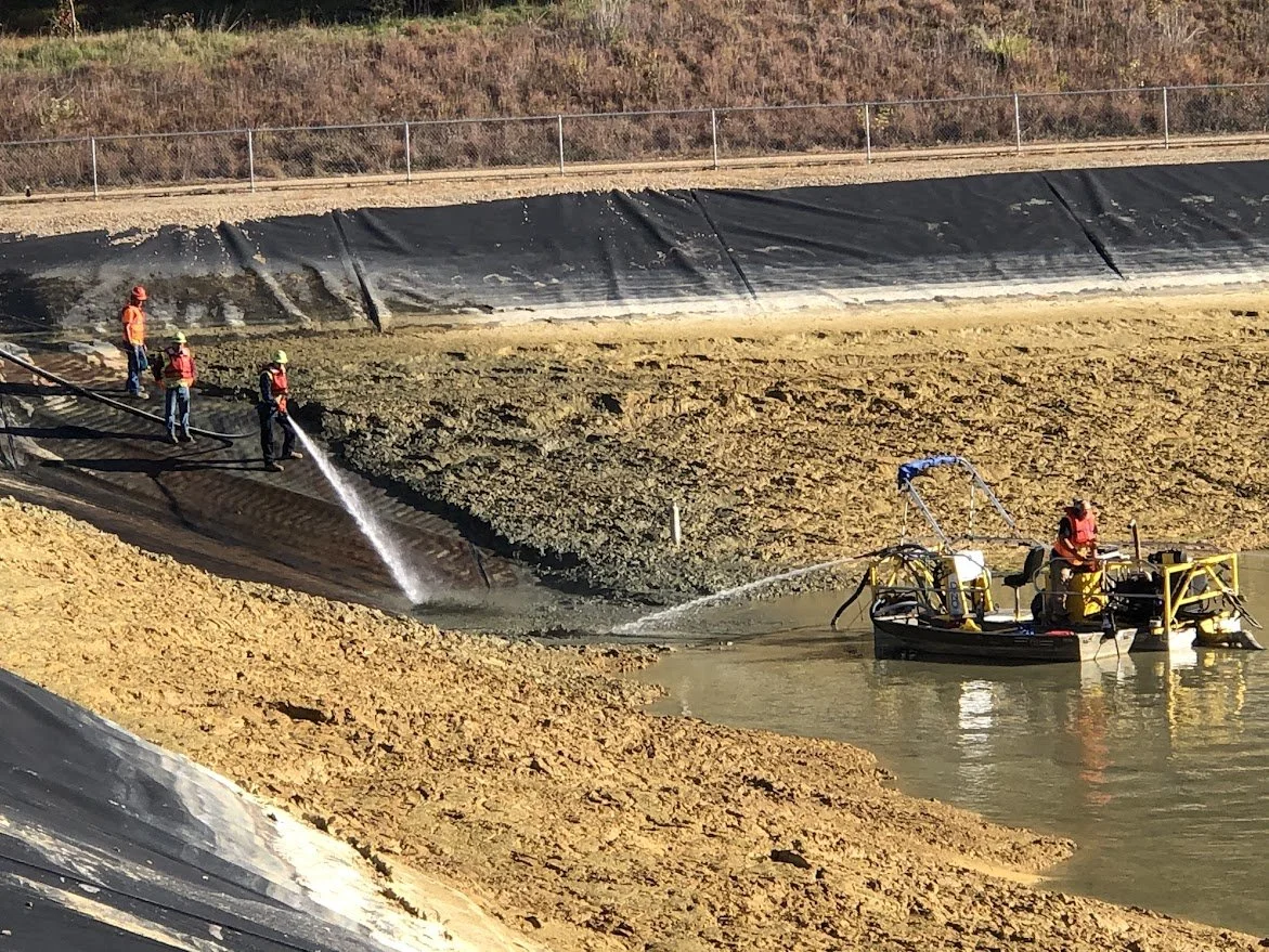 Construction workers cleaning a contaminated water detention basin using hoses, with some standing on the edge and others in a small boat.
