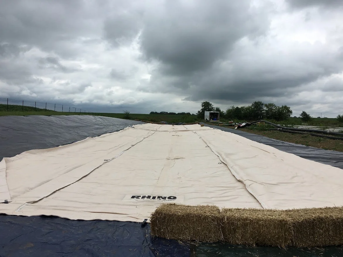 A large white tarp covering a section of outdoor ground, with black plastic around it and hay bales at one end, set in a rural area with cloudy skies and a trailer in the background.