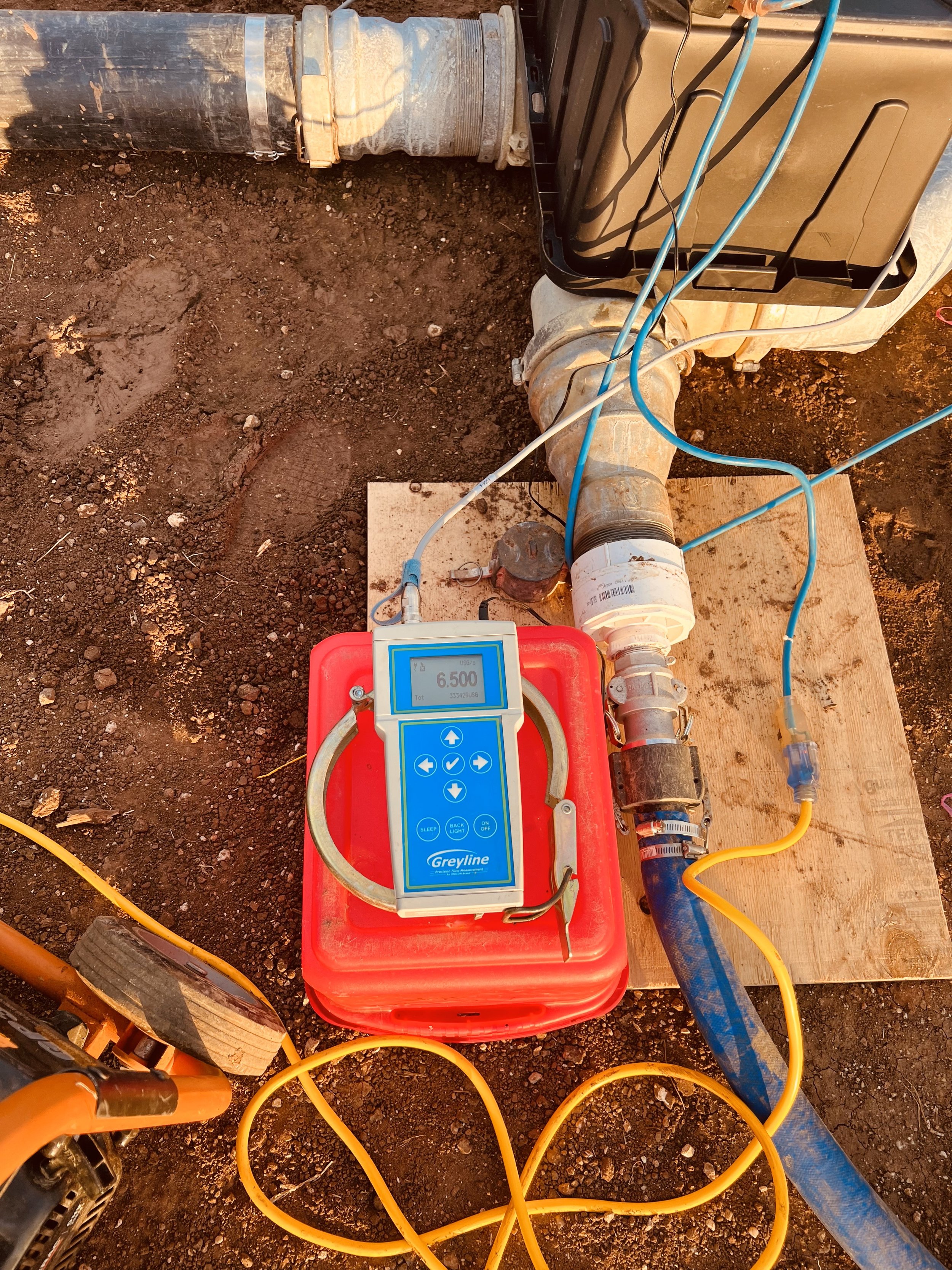 A setup for water testing or plumbing measurement outdoors, including a digital device, hoses, pipe fittings, and equipment on a wooden board with dirt ground.