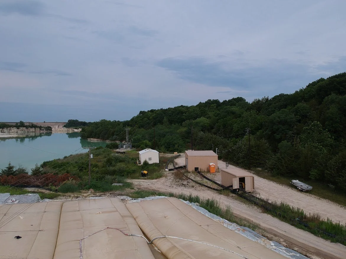 A view of a river with a forested hillside, dirt road, small buildings, and a trailer in a rural area under a cloudy sky.