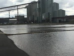 Industrial facility with large gray buildings and a conveyor system, located near a flooded area.