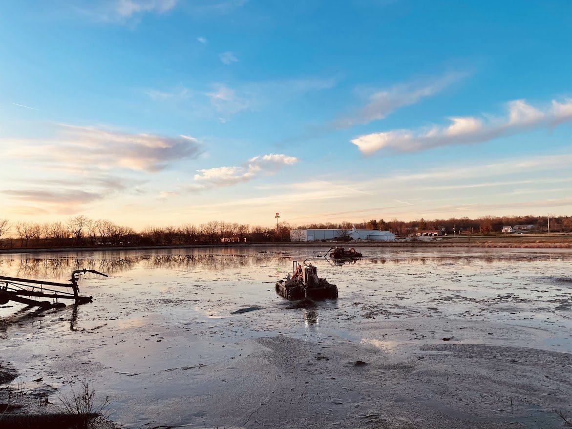 Body of water with two boats and one machine, clear sky with clouds at sunset.