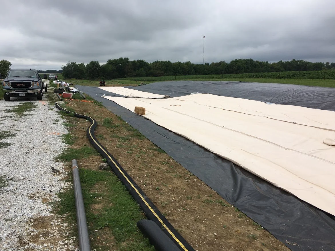 A large black and white irrigation tarpaulin spread across a field with vehicles parked on a gravel area nearby, under cloudy skies.