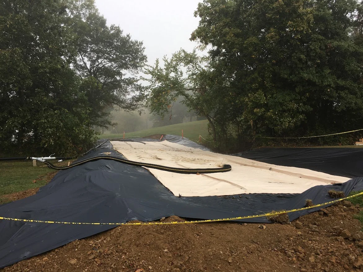 A construction site covered with black and white tarps, surrounded by yellow caution tape, and trees in the background.