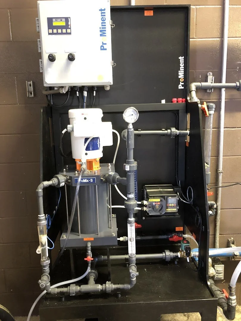 Industrial water treatment or filtration system with various pipes, gauges, and control units mounted on a black metal frame against a brick wall.
