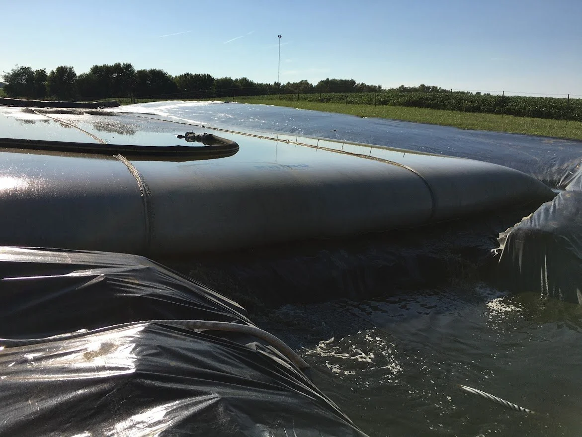 Close-up of large black plastic water storage tank, partially submerged in a body of water, with a rural landscape and blue sky in the background.
