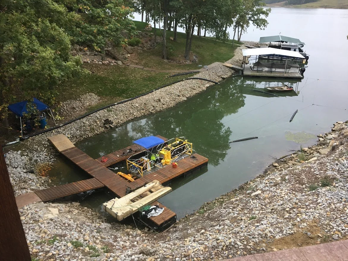 Dock and boats at a lakeside, with a small floating platform, two larger boats with canopies, and a small rowboat, surrounded by trees and rocky shoreline.