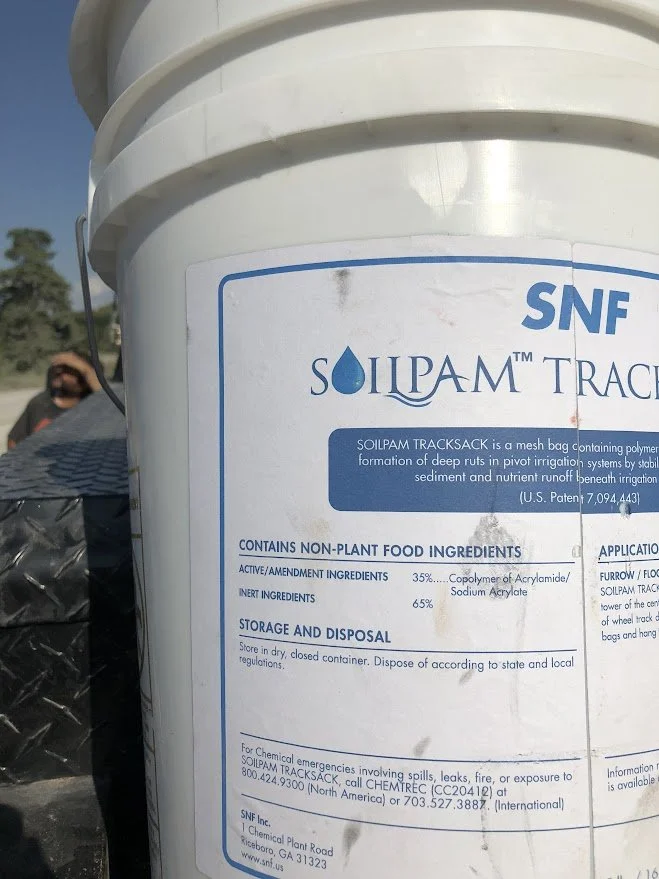 A white bucket labeled SOILPAM TRACKS containing a mesh bag product with non-plant food ingredients. Background shows a person wearing sunglasses and a black shirt, outdoors under a clear sky.