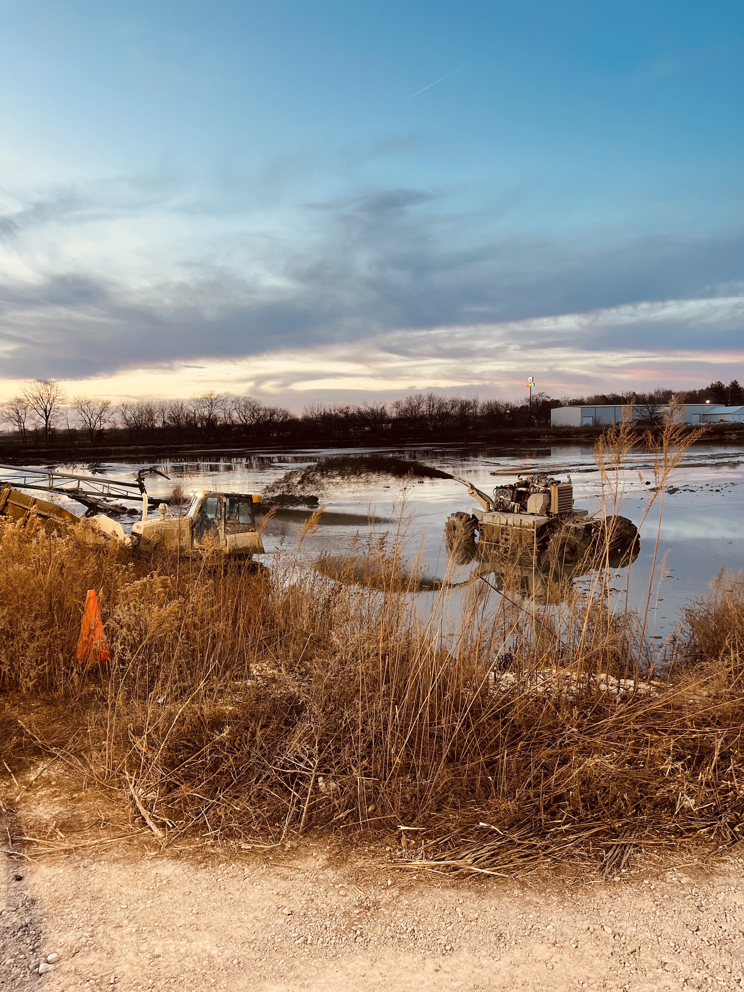 Construction equipment working in a flooded area with water, drying grass, and a cloudy sky during sunset or sunrise.
