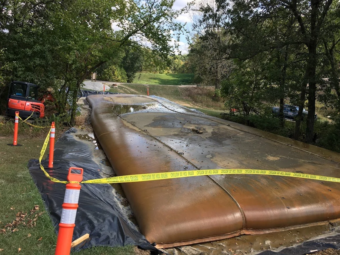 Heavy equipment and caution tape surrounding a deflated geotextile tube on a grassy area, with trees and a road in the background.