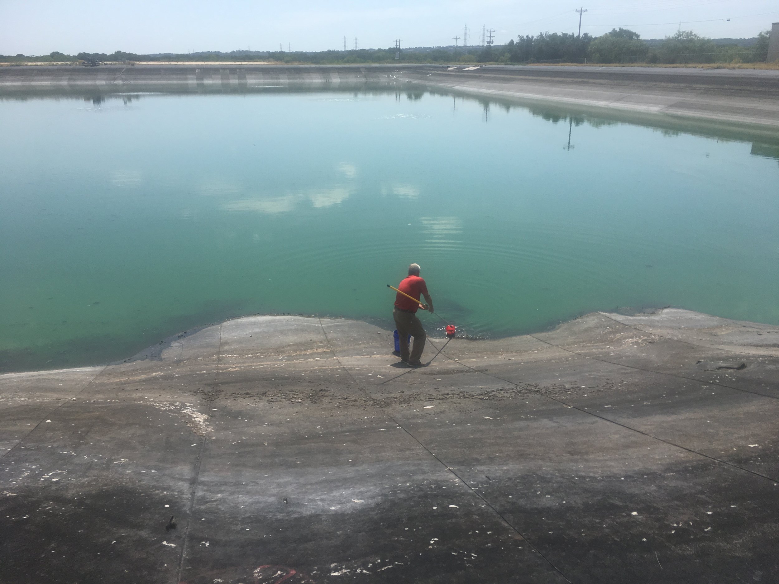 A person standing at the edge of a drained water reservoir, using a tool to inspect or work on the water of the reservoir.
