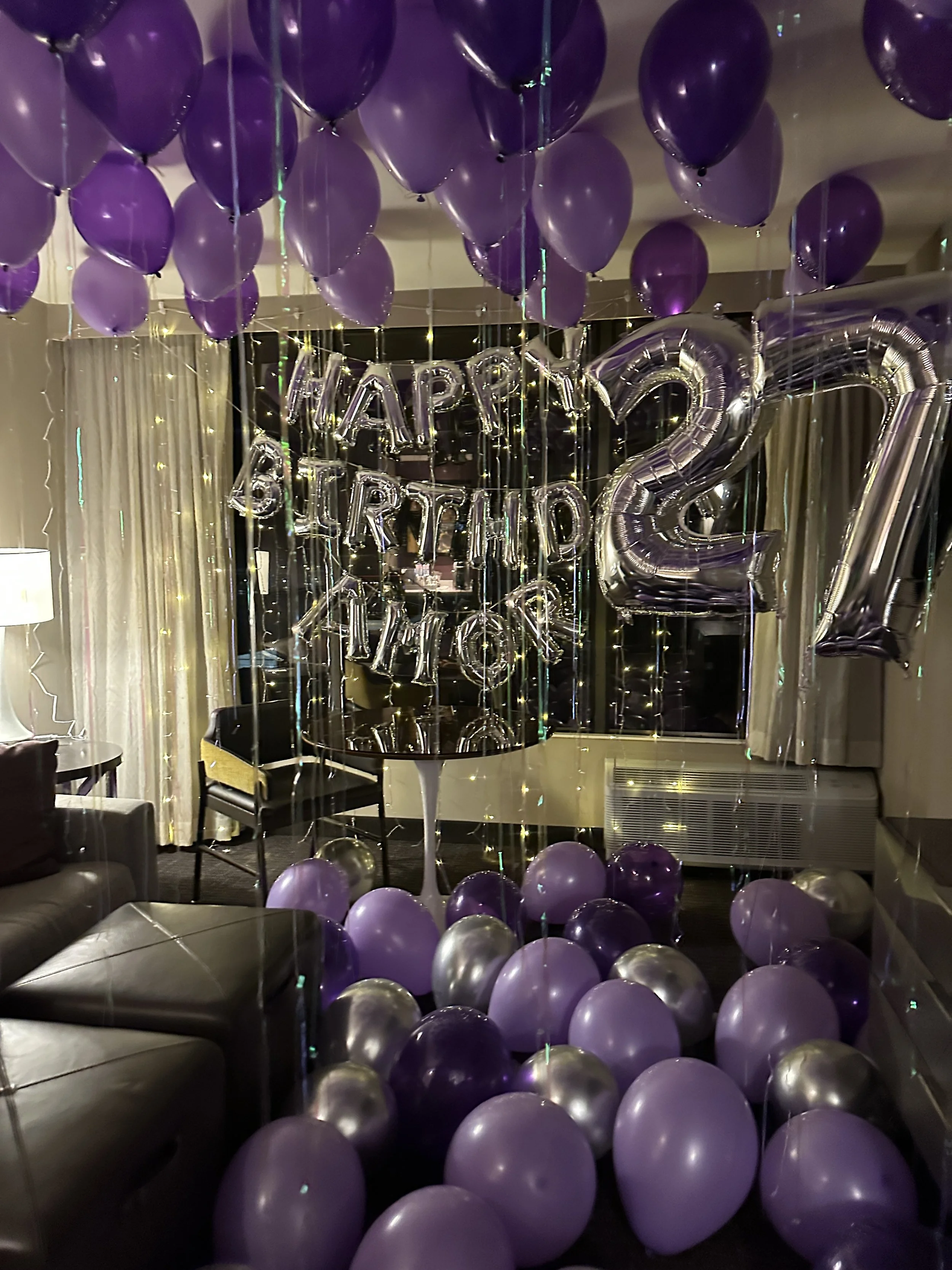 Hotel room decorated for a birthday celebration with gold and green balloons, including large numeral '32' balloons on the floor, a 'Happy Birthday Bryon' banner on the bed's headboard, and balloons with string decorations hanging from the ceiling.