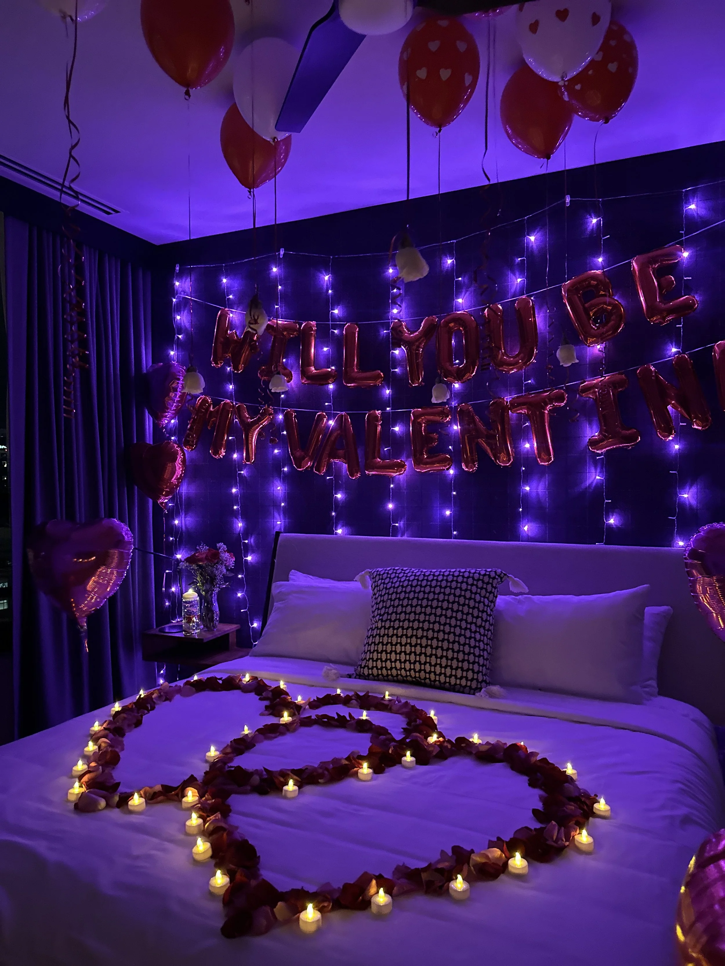 A decorated bedroom with purple string lights, valentine balloons, and candles forming a heart on the bed. The wall has a banner saying "Will You Be My Valentine".
