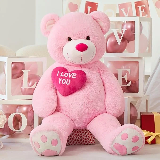 A large pink teddy bear holding a pink heart that says 'I Love You,' surrounded by Valentine's Day decorations including boxes of pink and rose-colored balloons and banners spelling 'LOVE.'