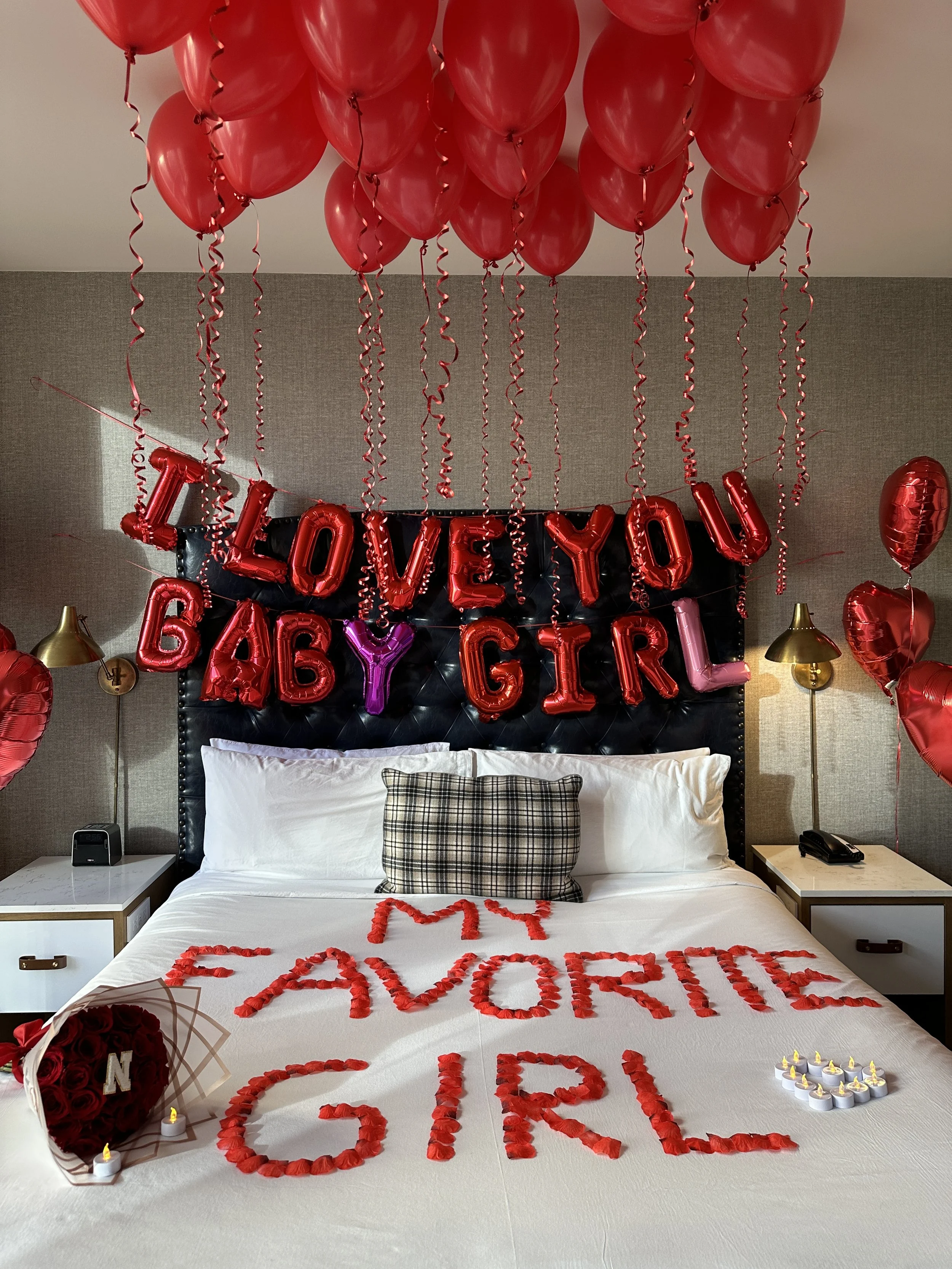 Valentine's Day bedroom decorated with red balloons and banners, with a message on the bed reading 'My Favorite Girl' made of red rose petals, a bouquet of roses, and candles.