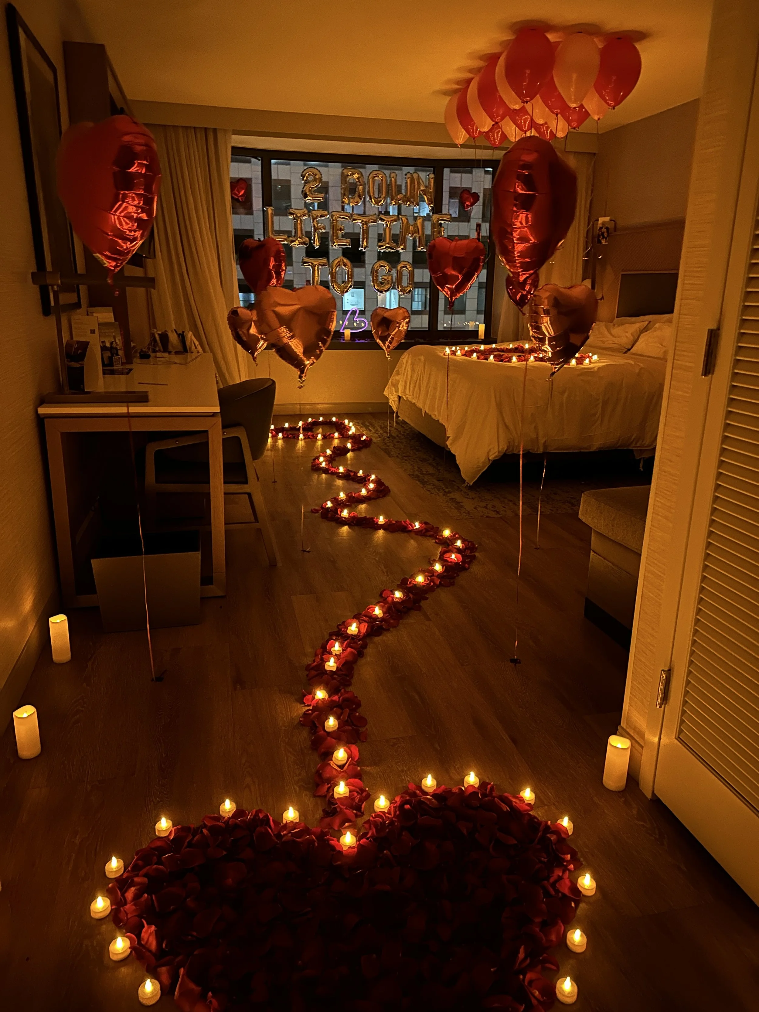 Decorated hotel room with green heart-shaped balloons, silver 'Happy Birthday Baby' balloons, tissue paper leaves arranged with tea light candles on the bed, and a cozy armchair beside the bed.