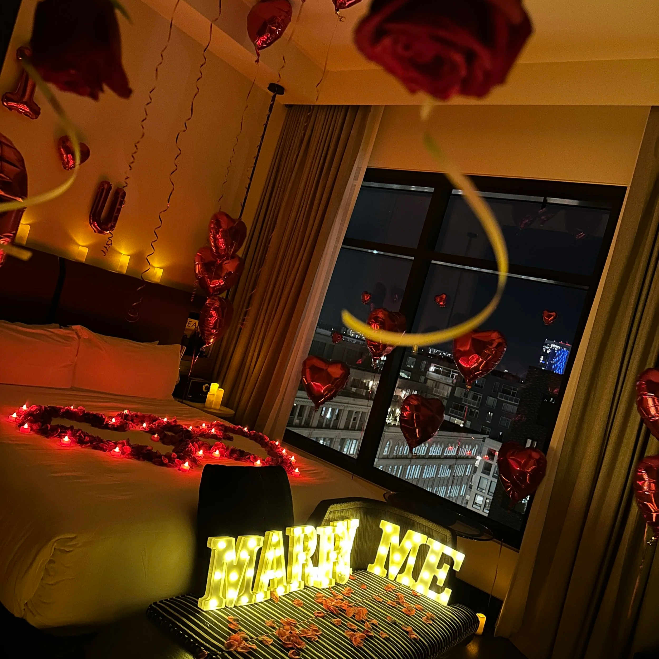 A hotel room decorated for a romantic occasion with purple and pink balloons, candles, and flower petals. A large heart shape made of rose petals and candles is on the bed. There are balloons hanging from the ceiling with the message "I LOVE YOU MORE," and purple heart-shaped balloons around the room.