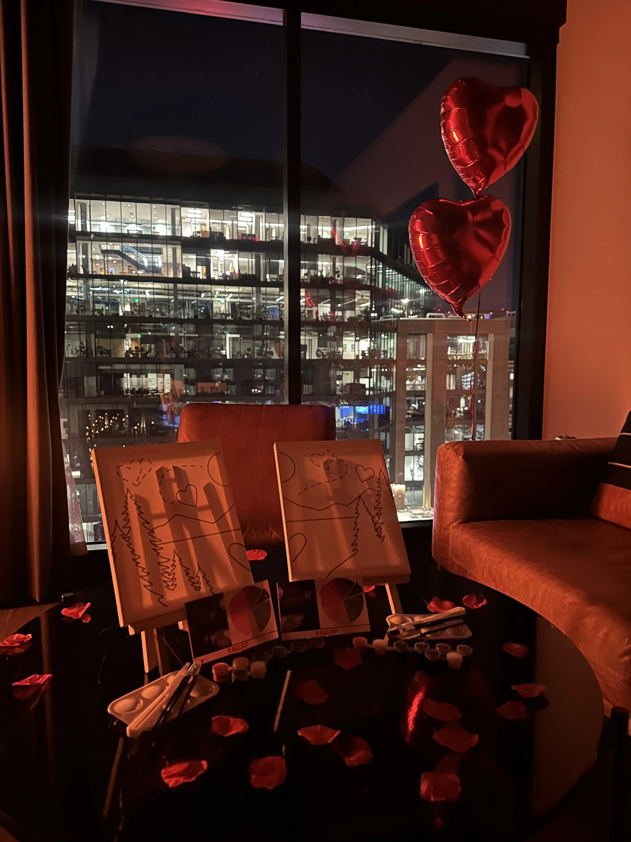 Valentine's Day setup with red heart-shaped balloons, rose petals on a table, and two paintings with hearts and trees, inside a room with a large window showing a cityscape at night.