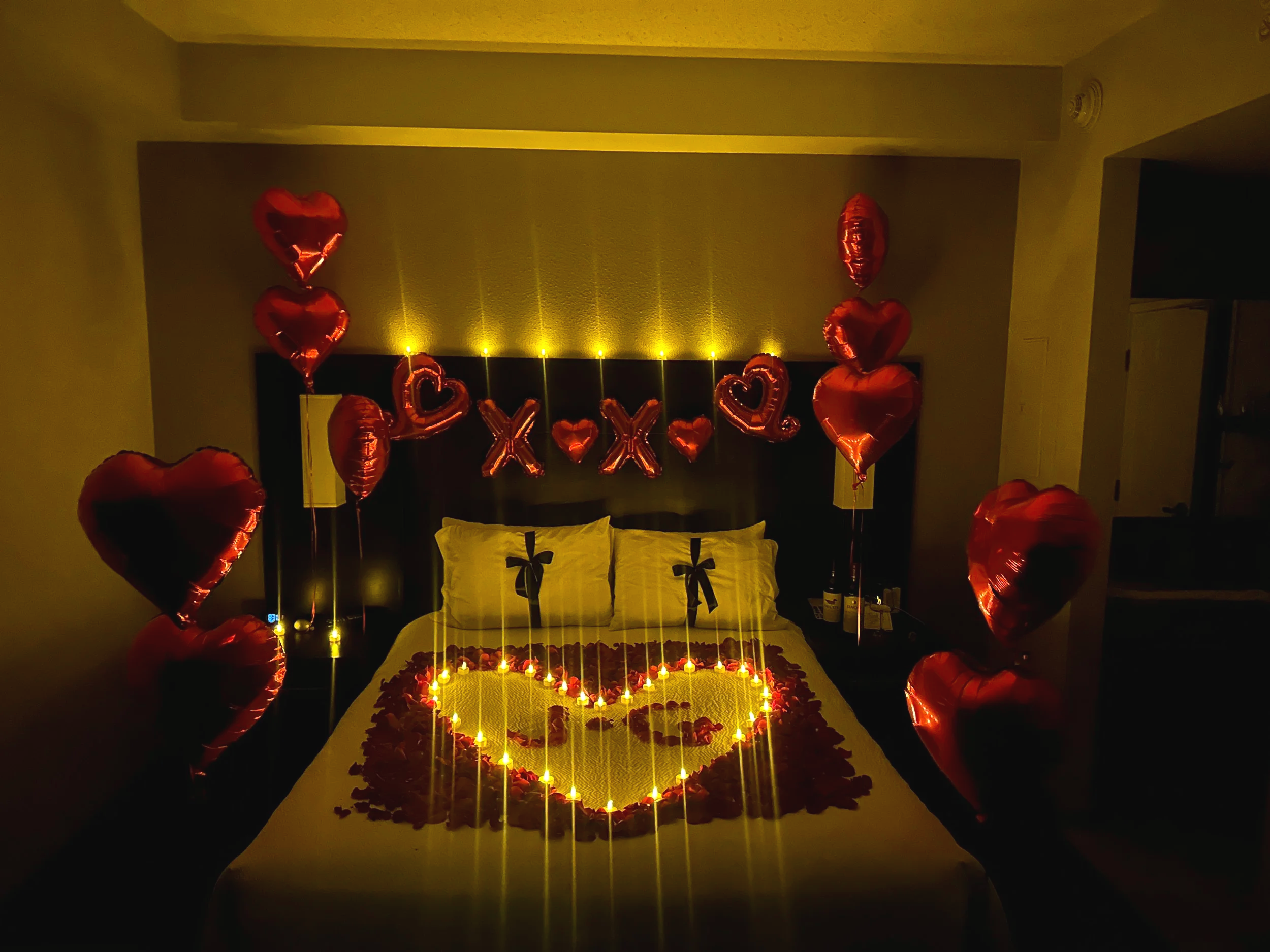 Decorated hotel room with pink and green balloons, a heart-shaped arrangement on the bed, and a 'Love You' balloon display on the wall.