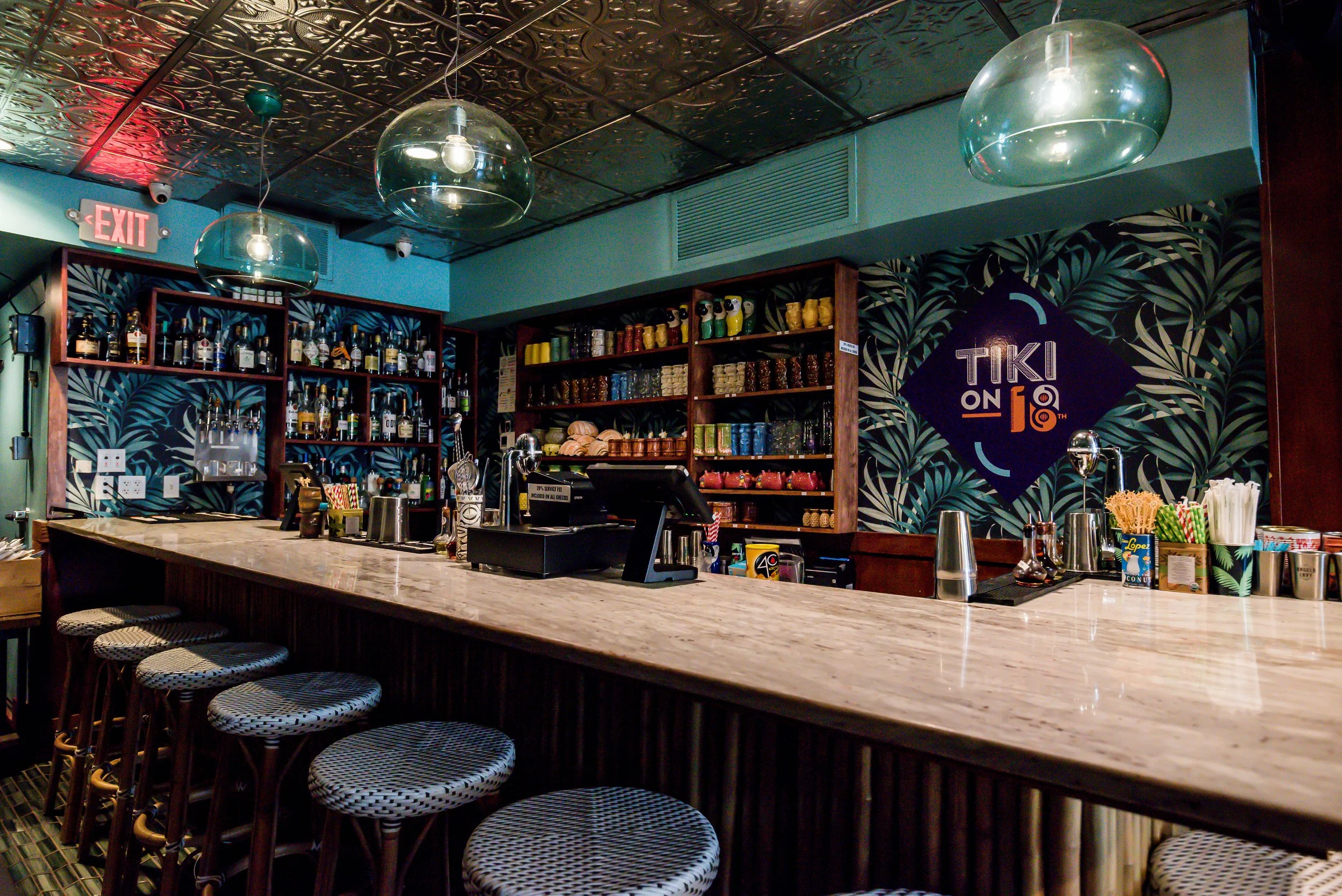 Cocktail bar with a marble counter, bar stools, shelves with bottles of alcohol, colorful cups, and tropical-themed wallpaper. There are modern pendant lights hanging from the ceiling.