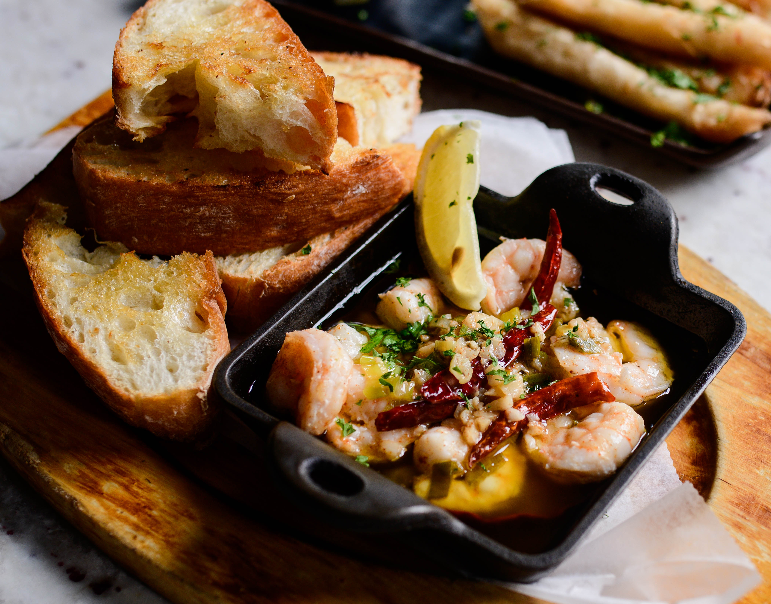 Gambas al Ajillo - with garlic, herbs, dried red chili peppers, served with sliced lemon and toasted bread on a wooden board.